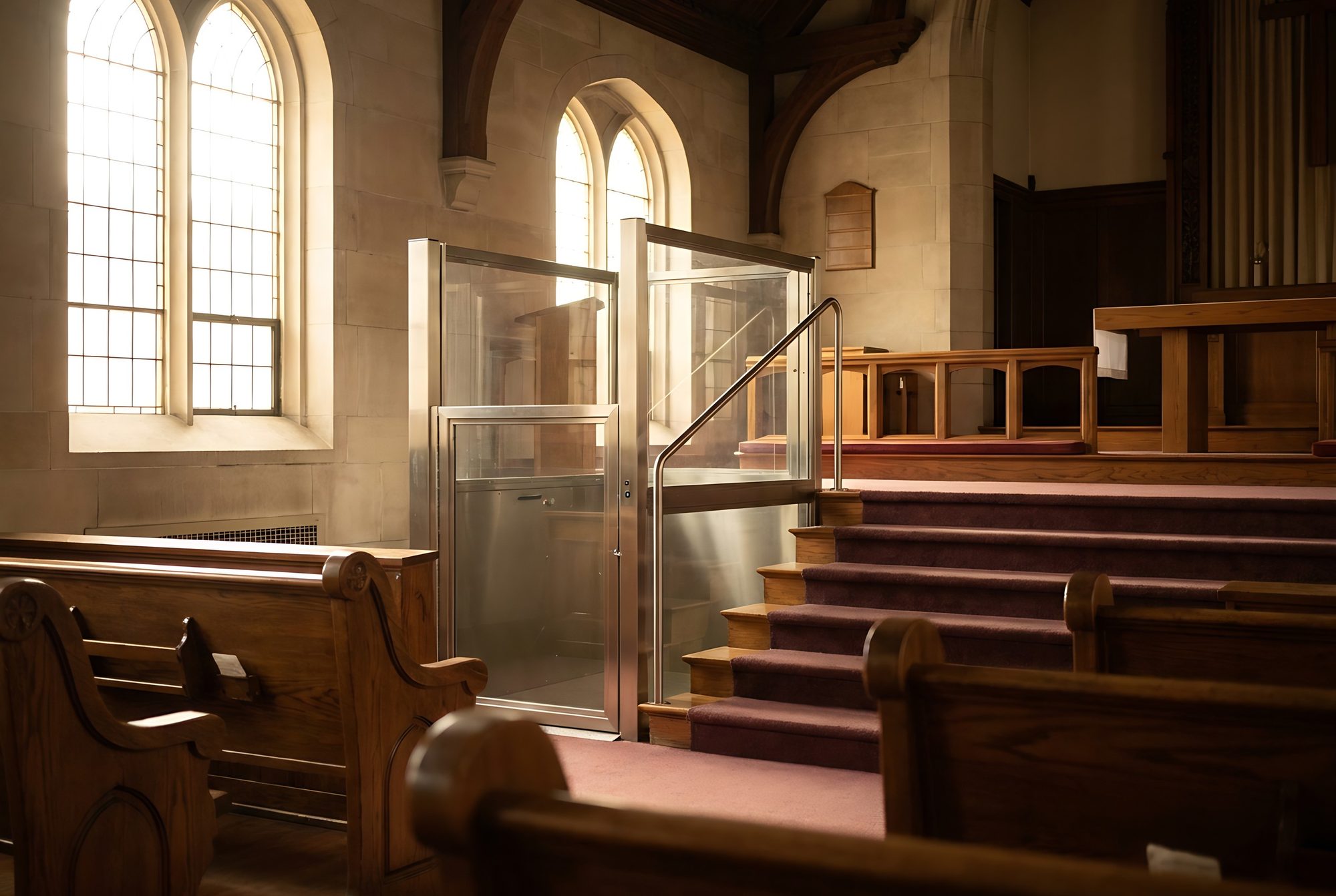 A glass wheelchair platform lift beside carpeted steps up to the platform in a stone church sanctuary, with wooden pews in the foreground and sunlight through arched windows