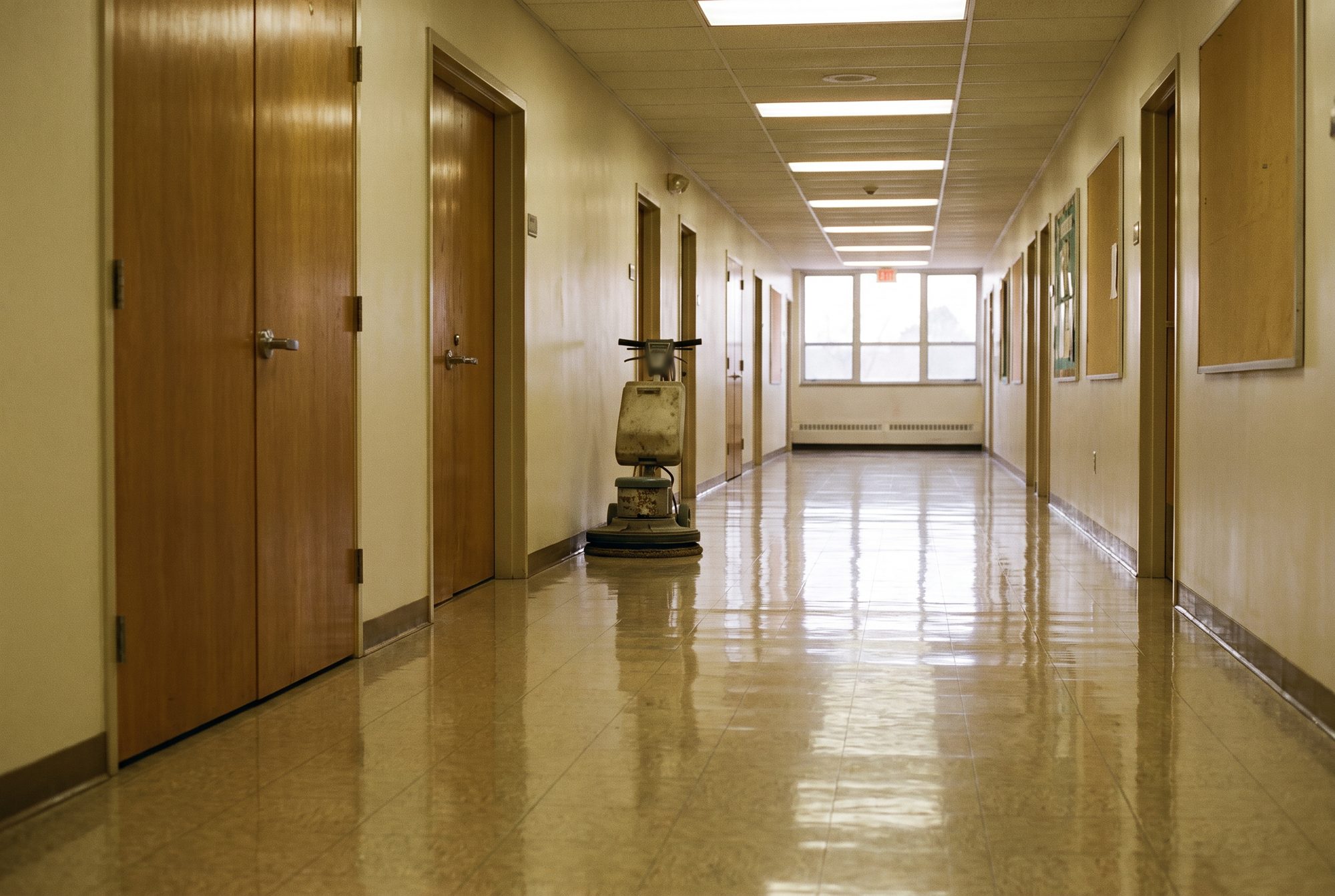 A long hallway in a church education wing, its glossy vinyl tile floor reflecting the ceiling lights, with a floor machine parked by the wall