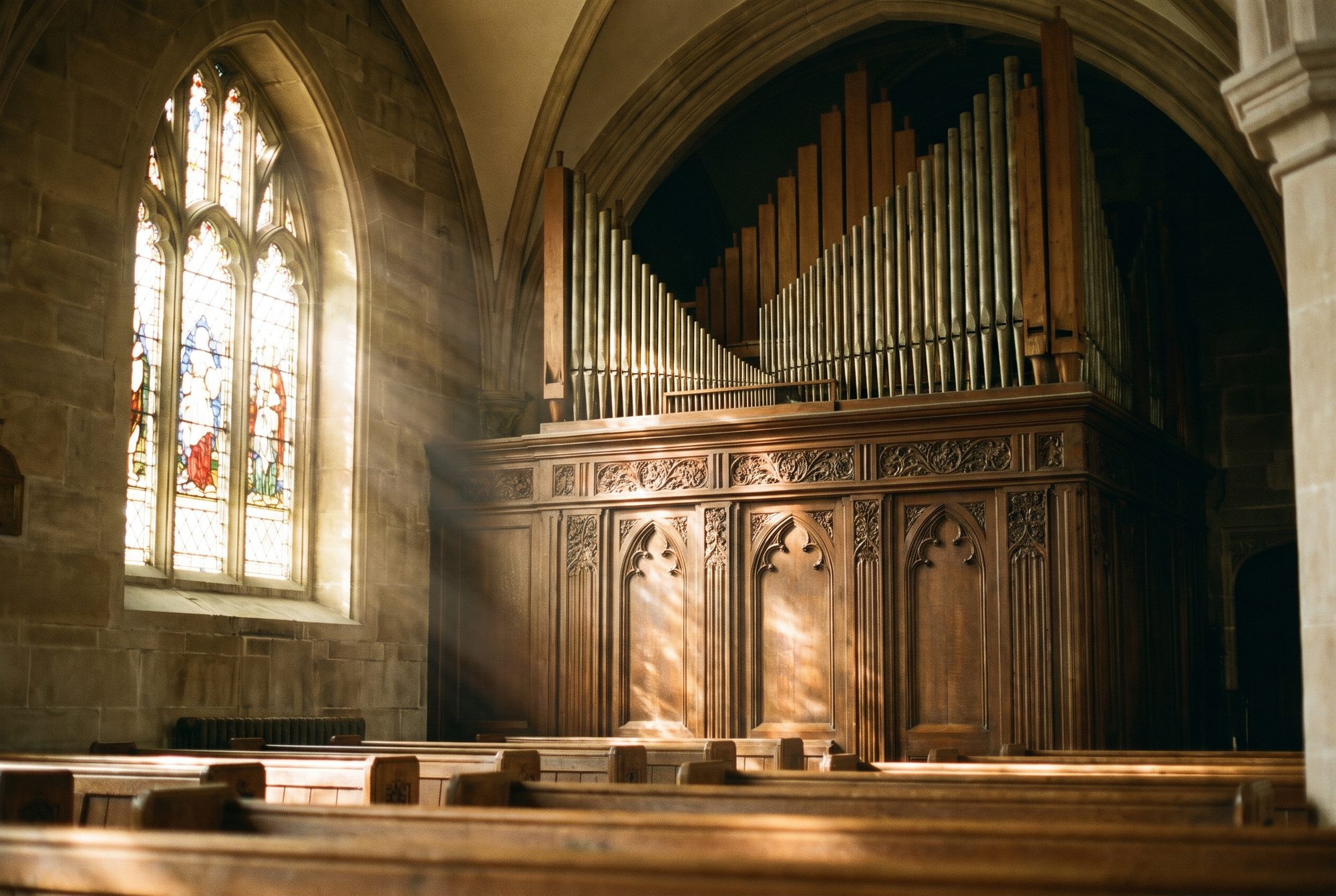 Pipe organ pipes rising above a carved wooden case in a stone church, lit by late-afternoon sun through a stained-glass window