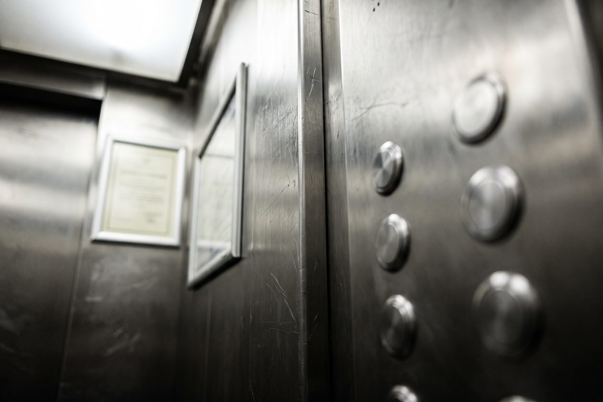 Inside an older elevator car with scratched steel walls, round buttons on the panel and a framed certificate on the wall beyond, out of focus